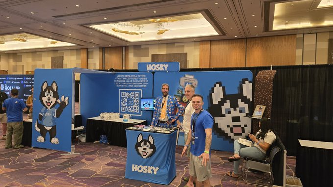Photo of a convention booth for 'HOSKY' cryptocurrency featuring a husky mascot, with three men posing at the display and a large pixel-art husky artwork on the wall.