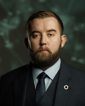 A professional headshot of a bearded man with short brown hair, wearing a dark suit, blue tie, and white shirt, with a circular pin on his lapel.