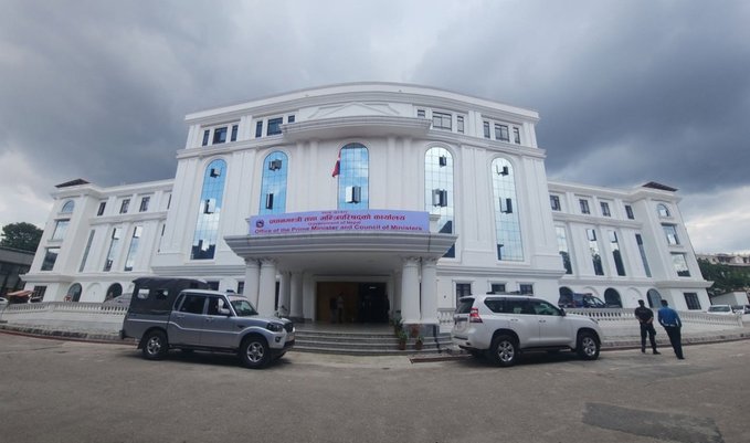 A grand white government building, identified by the banner above its entrance as the "Office of the Prime Minister and Council of Ministers," stands under a cloudy sky, with an SUV and a white Toyota Land Cruiser parked out front. The building's official designation directly relates to the post, which discusses efforts to route crypto aid into the PM Disaster Relief Fund following the floods.