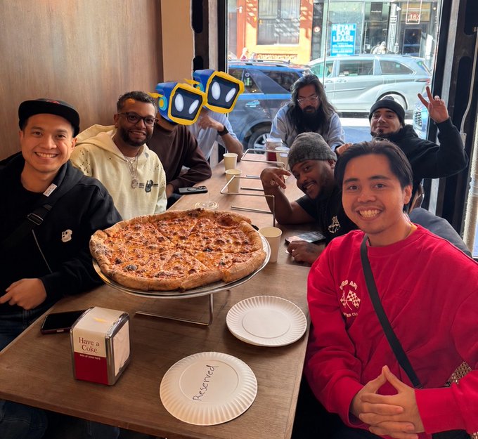 A group of men pose around a restaurant table with a large pizza, paper plates, cups, and phones; two faces are covered by yellow-eyed cartoon stickers.