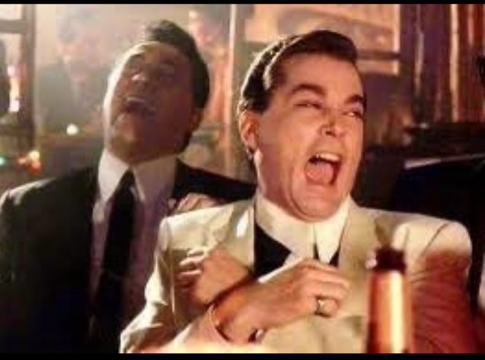 Two men in suits laughing hysterically in what appears to be a vintage movie scene, with one man wearing a cream-colored suit and a ring.