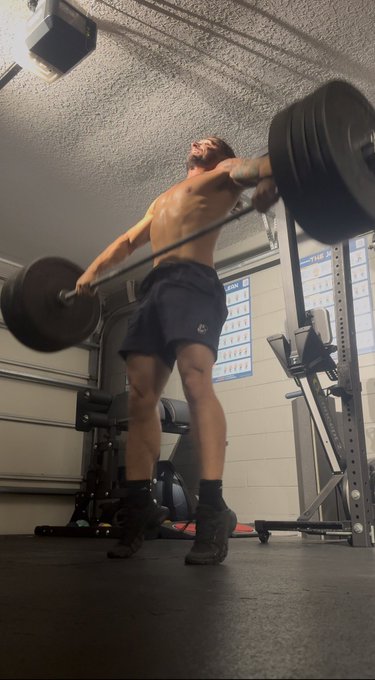 A shirtless man performs a heavy barbell lift in a garage gym, viewed from a low angle with the textured ceiling visible above.