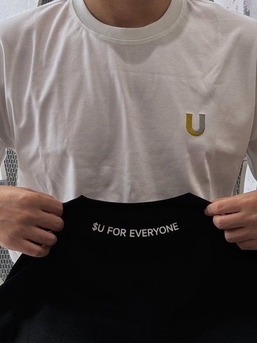 A person wearing a white t-shirt with a yellow 'U' logo holds up a black t-shirt displaying the text '$U FOR EVERYONE'.