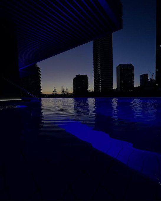 The image shows a large swimming pool with blue water, surrounded by tall buildings at dusk. The sky above the city skyline is dark, creating an atmosphere of tranquility as the sun sets. The photo captures the essence of a serene evening in the city, with the pool and its surroundings serving as a focal point for the viewer.