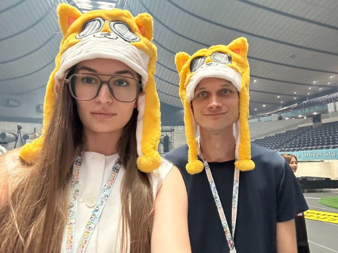 A young woman with glasses and a man pose for a selfie wearing matching yellow cat-ear trapper hats with aviator goggles, at what appears to be a stadium or convention venue with "University" visible on signage behind them. The post humorously suggests that even Ethereum co-founder Vitalik Buterin ("V God") cannot resist the allure of beauty, playing on his iconic cat-themed merchandise paired with a stylish companion.
