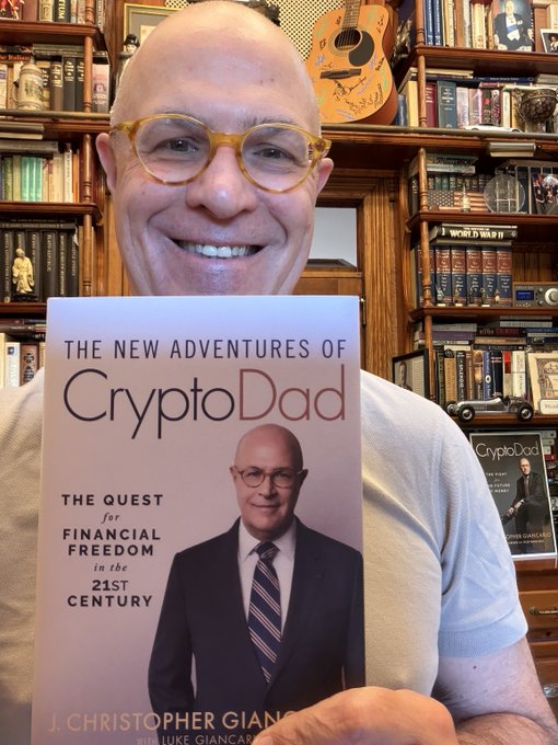 A smiling bald man wearing yellow-tinted glasses holds up his book "The New Adventures of CryptoDad: The Quest for Financial Freedom in the 21st Century" by J. Christopher Giancarlo, set against a backdrop of bookshelves, with another CryptoDad book visible behind him—fitting for a post highlighting the book's coverage of XRP and Ripple.