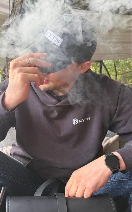 A man wearing a dark 'PYTH' branded hoodie and matching cap exhales vapor, wearing a smartwatch and a ring, seated outdoors.