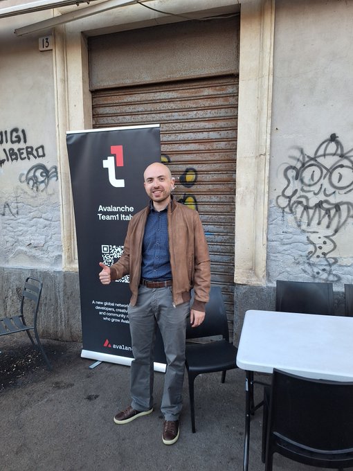 A bald man in a brown jacket and grey pants gives a thumbs-up in front of a black banner for the 'Avalanche Team1 Italy'. Graffiti is visible on the building behind him.