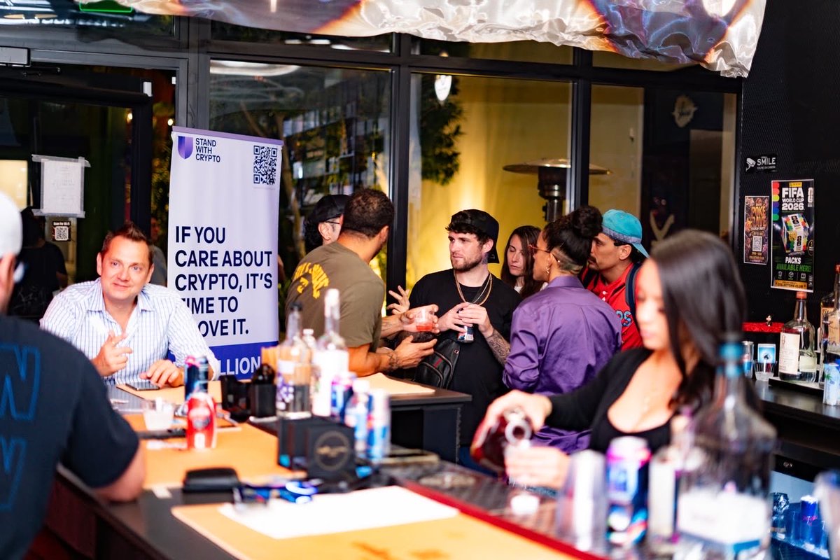 A group of people gathered at a bar during a 'Stand With Crypto' event, with a banner reading 'IF YOU CARE ABOUT CRYPTO, IT'S TIME TO PROVE IT' alongside a QR code.