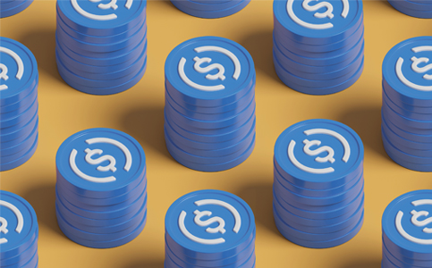 An arrangement of blue USDC cryptocurrency coins stacked in columns on a yellow/orange surface.
