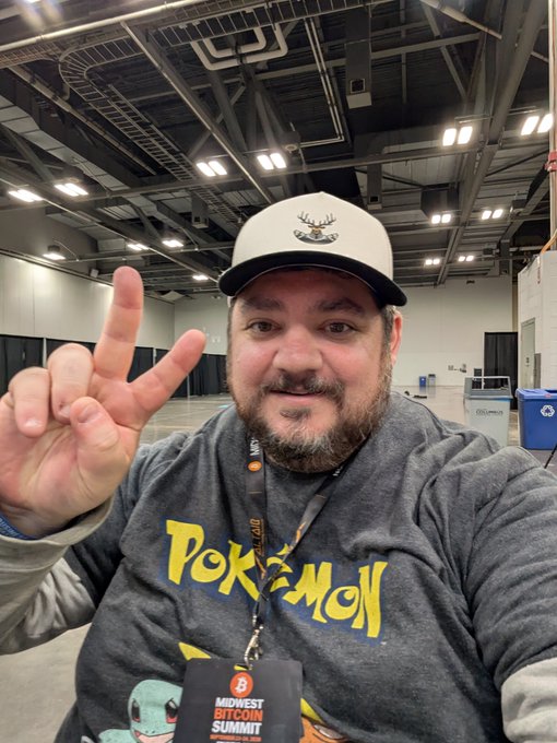 A bearded man wearing a Pokémon sweatshirt and a deer-logo cap poses with two fingers raised at the Midwest Bitcoin Summit, based on his event lanyard.