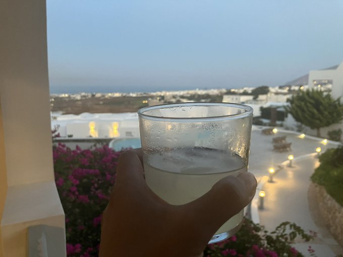 A hand holds a glass of cloudy liquid in the foreground, with a twilight view of white buildings, bougainvillea flowers, and the sea in the background.