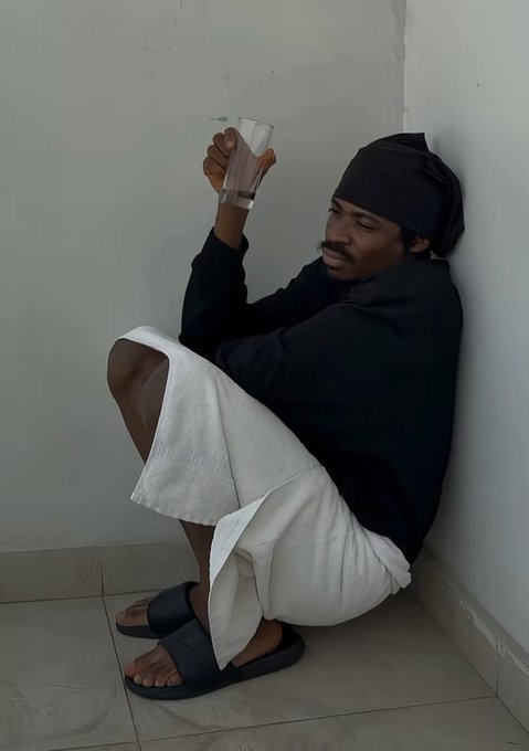 The image shows a man sitting down with his legs crossed, holding a glass of water while wearing slippers. The background appears to be a white wall or floor. There are no other people visible in the photo. The caption provided seems unrelated to the main subject and does not provide any context about the content of the image.