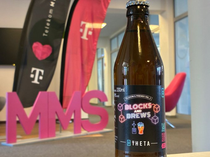 The image features a bottle of beer with "Blocks & Brews" written on it, along with a pink sign that says "MMS". The background shows a room with a TV screen and chairs, suggesting an event or gathering related to telecommunications and technology. The text in the image reads "We're excited to be joining Deutsche Telekom ( @mms_Blockchain ) later this week at their event at @BerBlockWeek They've created 50 custom Telekom & Friends: Blocks & Brews beer bottles featuring Theta branding, which will be available exclusively at the event."