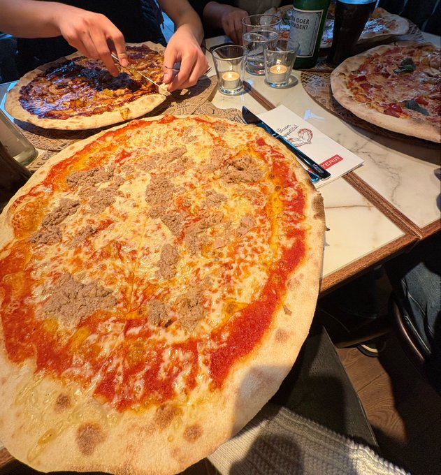 A close-up photo of a large pizza with tuna and tomato sauce on a table at a restaurant, with two other partially visible pizzas and lit candles nearby.