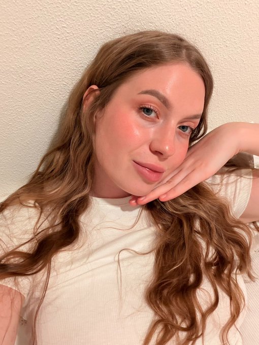 A close-up selfie shows a young woman with long wavy reddish-brown hair, blue eyes, and dewy makeup, resting her face against her hand.