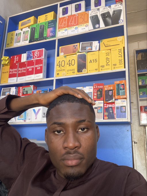 A man takes a selfie in front of shelves displaying various smartphone boxes, including brands like Redmi A3, Redmi A5, Hot 40, Hot 50, Pop 10, and Tecno.