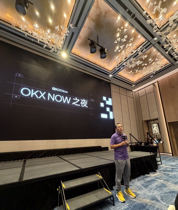 A man in a purple polo shirt and yellow sneakers stands on a stage speaking into a microphone, with a large screen behind him displaying 'OKX NOW 之夜' (OKX NOW Night).