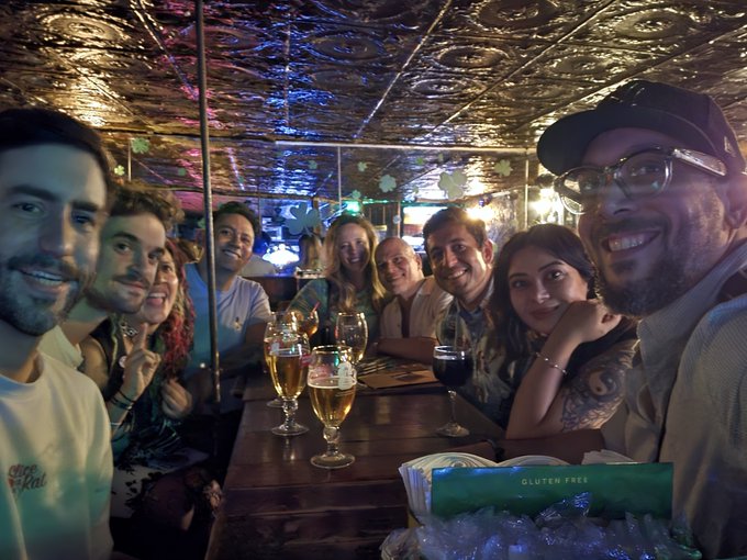 A group of seven friends poses together at a dimly lit bar with a decorative metallic ceiling, with beer and wine glasses on the table and a "Gluten Free" menu visible.