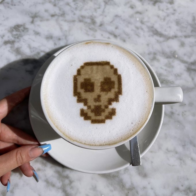 A hand with blue nail polish holds a cappuccino on a marble table, featuring pixel art of a skull design made with cocoa powder in the foam.