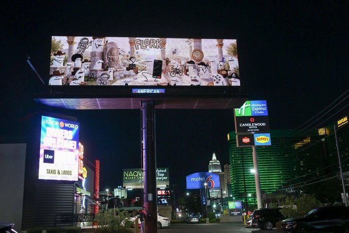 A brightly illuminated digital billboard on the Las Vegas Strip displays an advertisement for "$FLORK," featuring cartoon-style meme characters alongside a smartphone graphic, towering above the street at night. Surrounding signage for Hollywood Car Wash, Uber "Mystery Vehicles" taxis/limos, the Holiday Inn Club Vacations, and Candlewood Suites, as well as the distant MGM building, confirms the location matches the post's claim that "$FLORK is live on the Las Vegas Strip for a full week, right next to the MGM Casino."