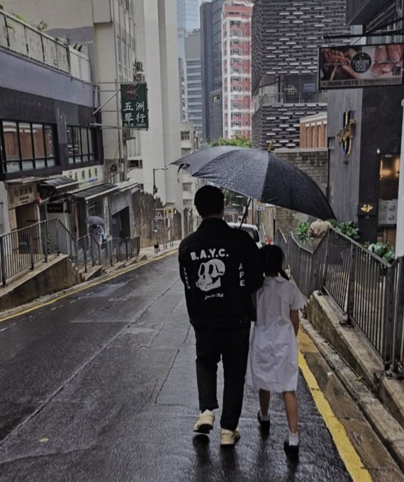 A photograph of two people walking down a rainy city street under a black umbrella; one person wears a black jacket with 'B.A.Y.C' and a skull logo on the back.