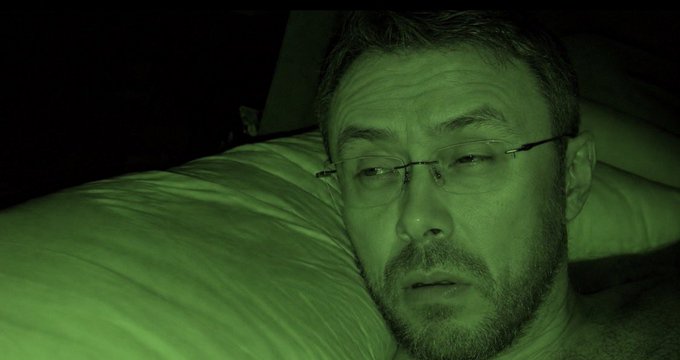 A close-up, green-tinted photograph of a bearded man with glasses lying in bed, looking at the camera with a tired expression.
