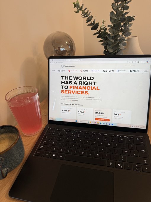 A workspace setup with an open laptop displaying 'THE WORLD HAS A RIGHT TO FINANCIAL SERVICES' from 'Bank for Humanity' showing 301.4M donors, alongside a pink drink and ceramic mug on a wooden desk.