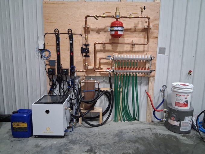 Industrial plumbing equipment setup mounted on a plywood board, featuring copper pipes, a red expansion tank, green PEX tubing manifolds, and various pumps and containers including ones labeled 'FRECOIL' and 'CRYO TEK-100'.