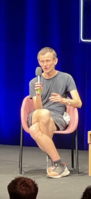 The image shows a man, identified as Vitalik Buterin, sitting casually on a pink chair on stage while holding a microphone and speaking at an event, matching the viral post about his distinctive seated posture. He is wearing a grey t-shirt, shorts, and sneakers, with a blue curtain backdrop behind him.