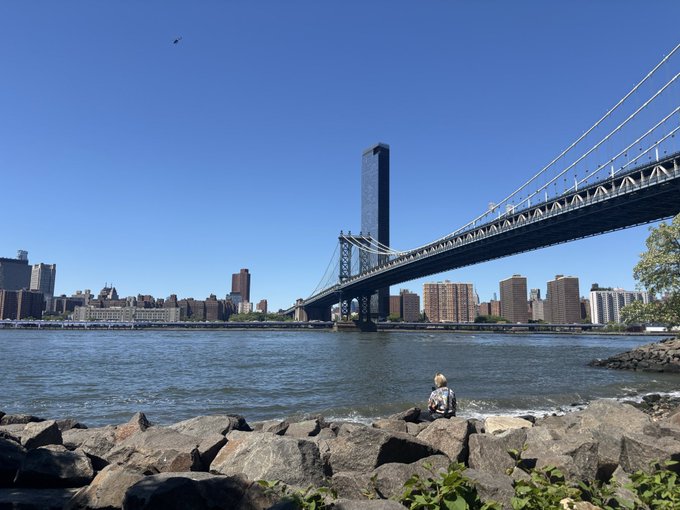 A scenic view of the Manhattan Bridge spanning the East River in New York City, with a person sitting on the rocky shoreline looking out at the water and skyline on a clear day.