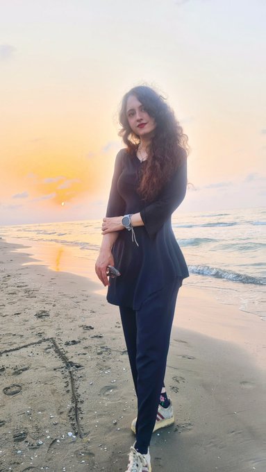 A portrait of a woman with long curly hair standing on a beach at sunset, wearing a black top and pants, with the ocean and sky in the background.