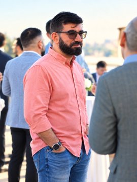 A bearded man wearing sunglasses and a pink button-up shirt stands outdoors at what appears to be a social gathering, with his hands in his pockets.