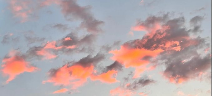 Pink and orange clouds are illuminated by the setting or rising sun against a light blue sky.