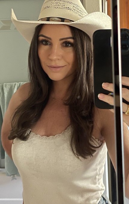 A woman takes a mirror selfie wearing a white cowboy hat and a cream-colored ribbed tank top with lace trim.