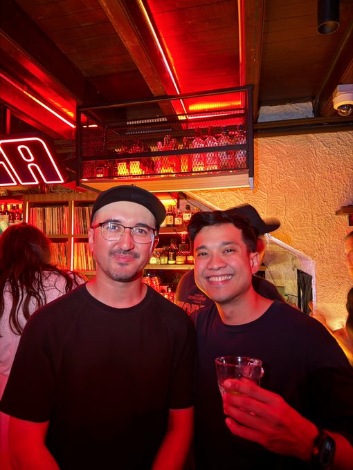 Two men smiling at a bar with red neon lighting, one wearing glasses and a cap, the other holding a drink.