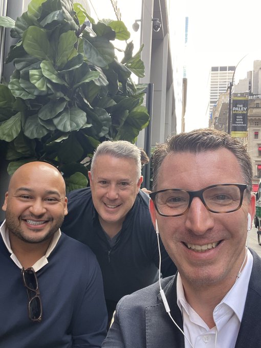 A selfie of three men outdoors, with the man on the right wearing earphones, taken near a large potted plant on a city street.