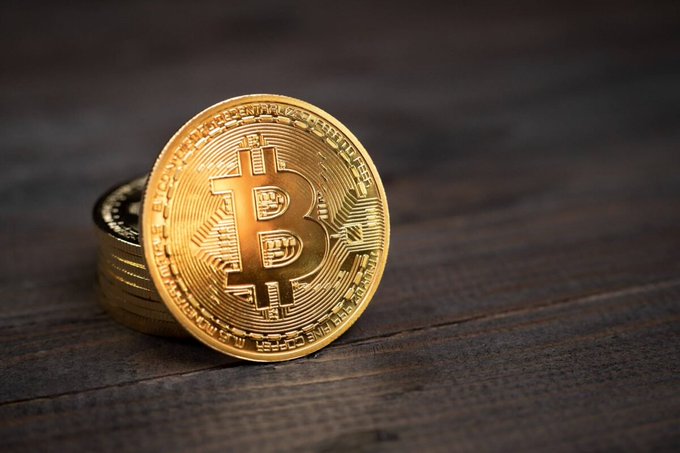 A close-up photograph of stacked golden Bitcoin tokens resting on a dark wooden surface.