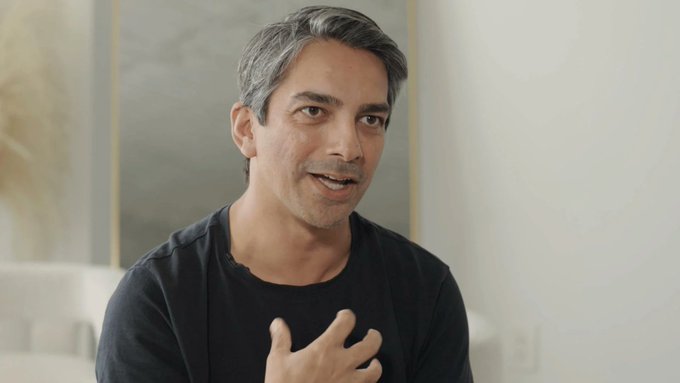 The image features a man wearing a black shirt with his hands up to his chest, possibly gesturing or making a point during a conversation. The background of the image shows a white wall, which contrasts with the gray floor beneath him. There is also an additional person partially visible in the top left corner of the frame.