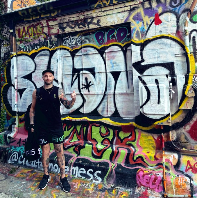 A tattooed man in a black tank top and shorts gives a thumbs-up while standing in front of a wall heavily covered in colorful graffiti featuring the word 'BIRD' and various tags.