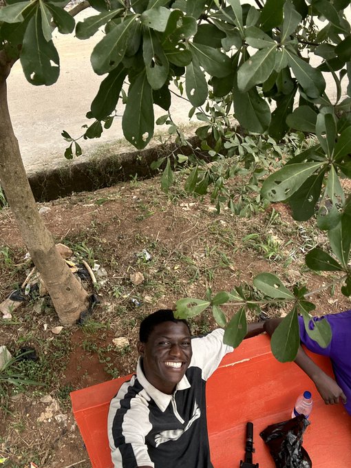 A smiling Black man in a black and white shirt relaxing under a leafy tree, leaning on an orange surface where a toy rifle and bottle are visible.