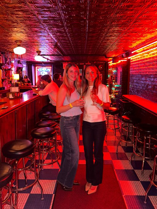 Two women smile and pose with drinks inside a neon-lit bar featuring red lighting, a tin ceiling, checkered floor, and bar stools.