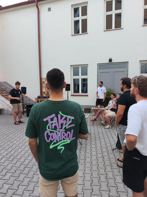 Casual outdoor gathering with several people, focusing on a man wearing a green t-shirt with the text "TAKE CONTROL" in purple and green graffiti-style lettering.