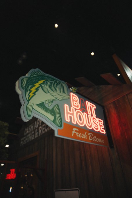 A neon-lit "Bait House - Fresh Bitcoin" sign featuring a large bass fish glows against a dark wooden storefront, cleverly blending traditional fishing imagery with cryptocurrency references for CashApp's Bitcoin-themed attraction at ComplexCon.