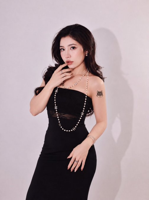The image features a woman wearing a black dress with pearls around her neck. She has a tattoo of a cat on her shoulder and is posing for a picture against a white background, which highlights her attire and the tattoo. The caption suggests that she might be interested in getting a tattoo or has recently gotten one done.