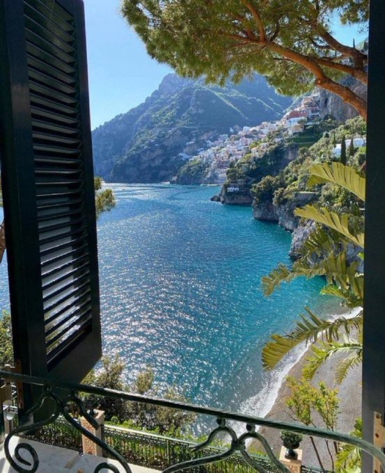 A scenic view through an open window with dark shutters reveals a bright blue body of water bordered by steep, sunlit mountains and a coastal town.