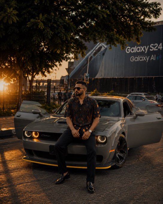 A man in sunglasses and a patterned shirt leans against a grey Dodge Challenger with its doors open, in front of a billboard advertising the 'Galaxy S24 Galaxy AI'.