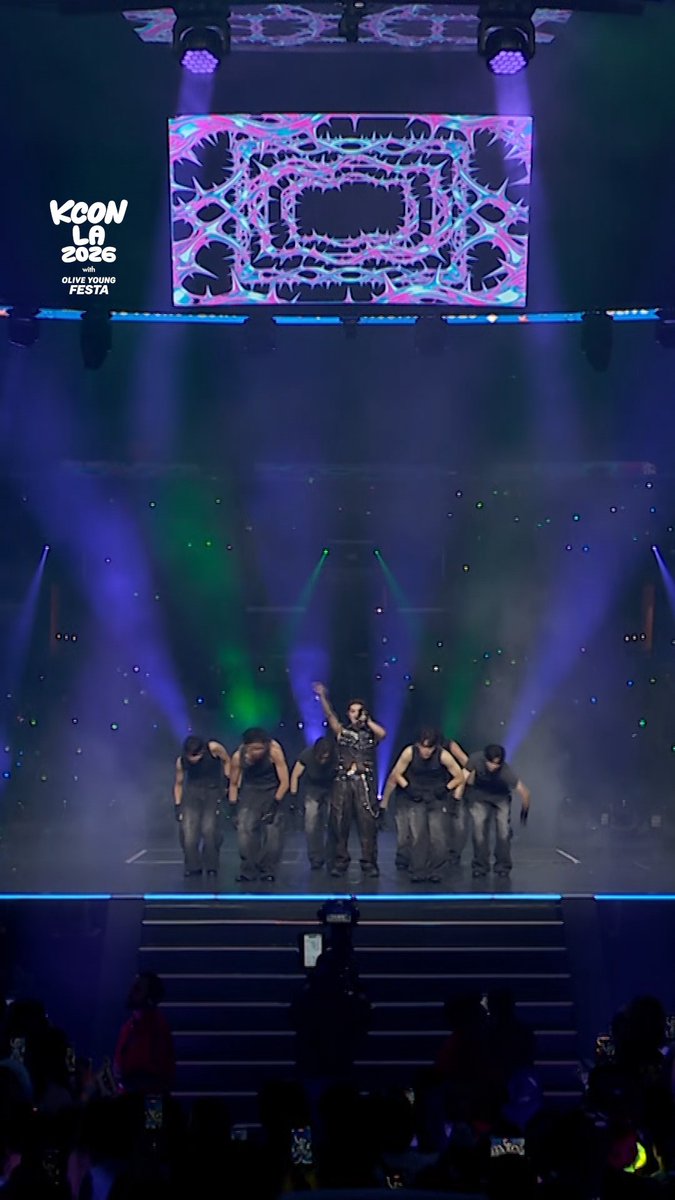 A K-pop performance at KCON LA 2026 with Olive Young Festa, showing artists on stage in dark outfits under purple and blue stage lighting, with a large psychedelic screen behind them and an audience holding green light sticks.