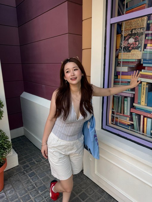 A young woman posing next to a colorful window display featuring a 'Hawaii Cook Book,' wearing a light top, white shorts, and red shoes, with a blue tote bag.