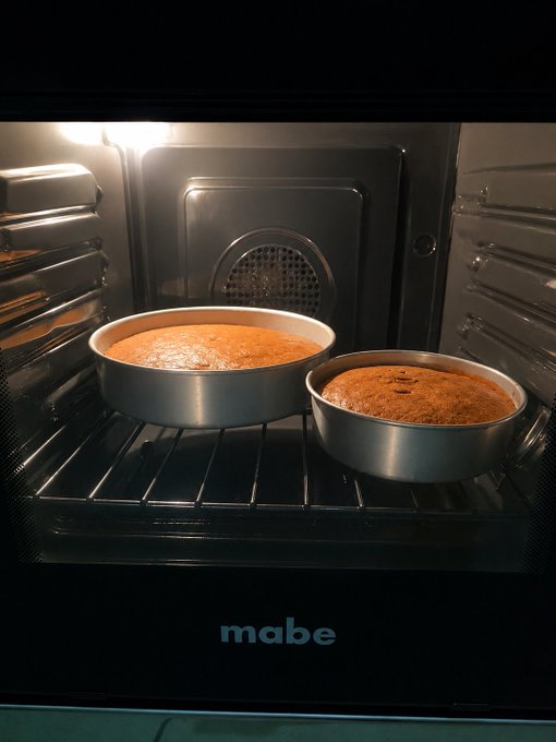 Two round cakes baking in round metal pans on a rack inside a lit Mabe brand oven.